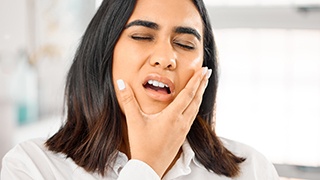 Woman with dark hair closing eyes holding hand around jaw in pain